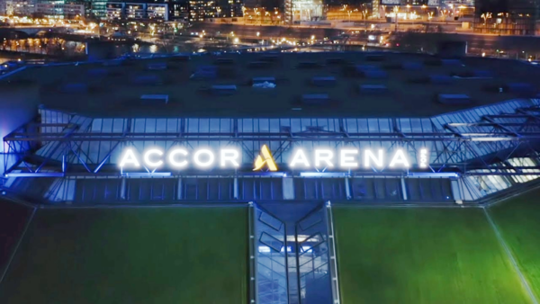 Nächtliche Luftaufnahme der Außenansicht der Accor Arena mit beleuchtetem Logo und Lichtern der Stadt im Hintergrund.