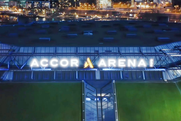 Nächtliche Luftaufnahme der Außenansicht der Accor Arena mit beleuchtetem Logo und Lichtern der Stadt im Hintergrund.