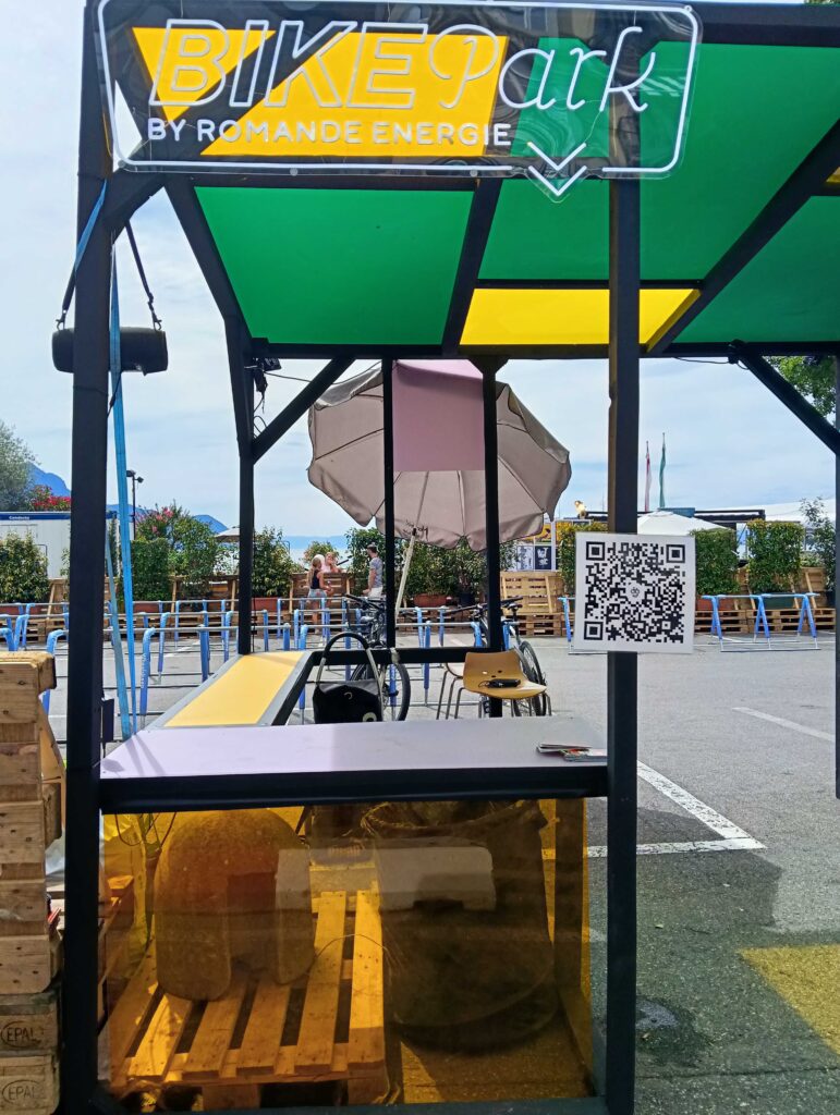Outdoor-Fahrradpark-Kiosk mit einem Neon-„BIKEpark“-Schild, grünen und gelben Paneelen, Holzpaletten und einem nahegelegenen QR-Code-Schild; Menschen und Fahrräder im Hintergrund sichtbar.