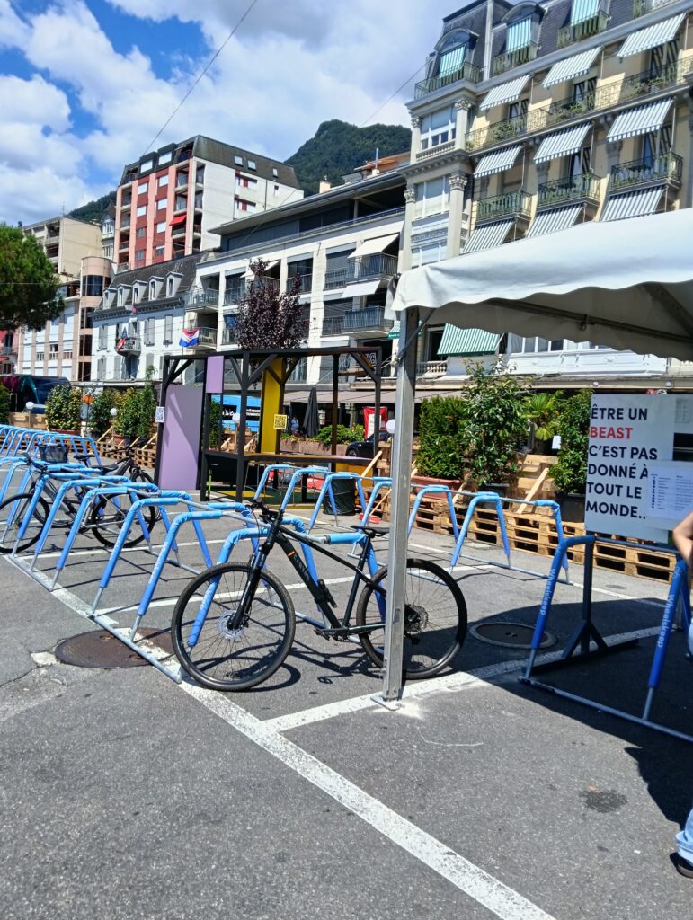 Reihe leuchtend blauer Fahrräder, die in einem Fahrradständer auf einem sonnigen städtischen Platz geparkt sind, mit Wohngebäuden und einem Hügel im Hintergrund